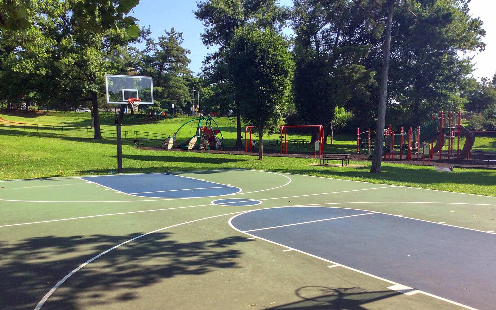Outdoor basketball court with painted lines in a park, with playground equipment and trees in the background on a sunny day.