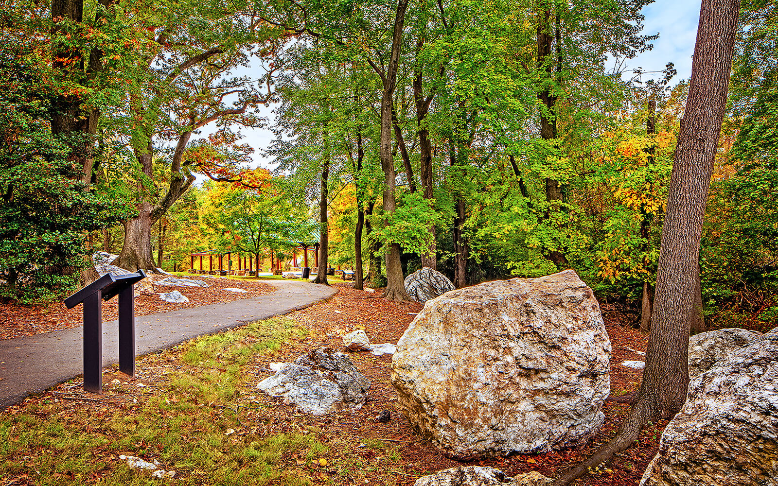 A paved pathway curves through a wooded area with autumn foliage, large rocks, and an informational sign on the left.