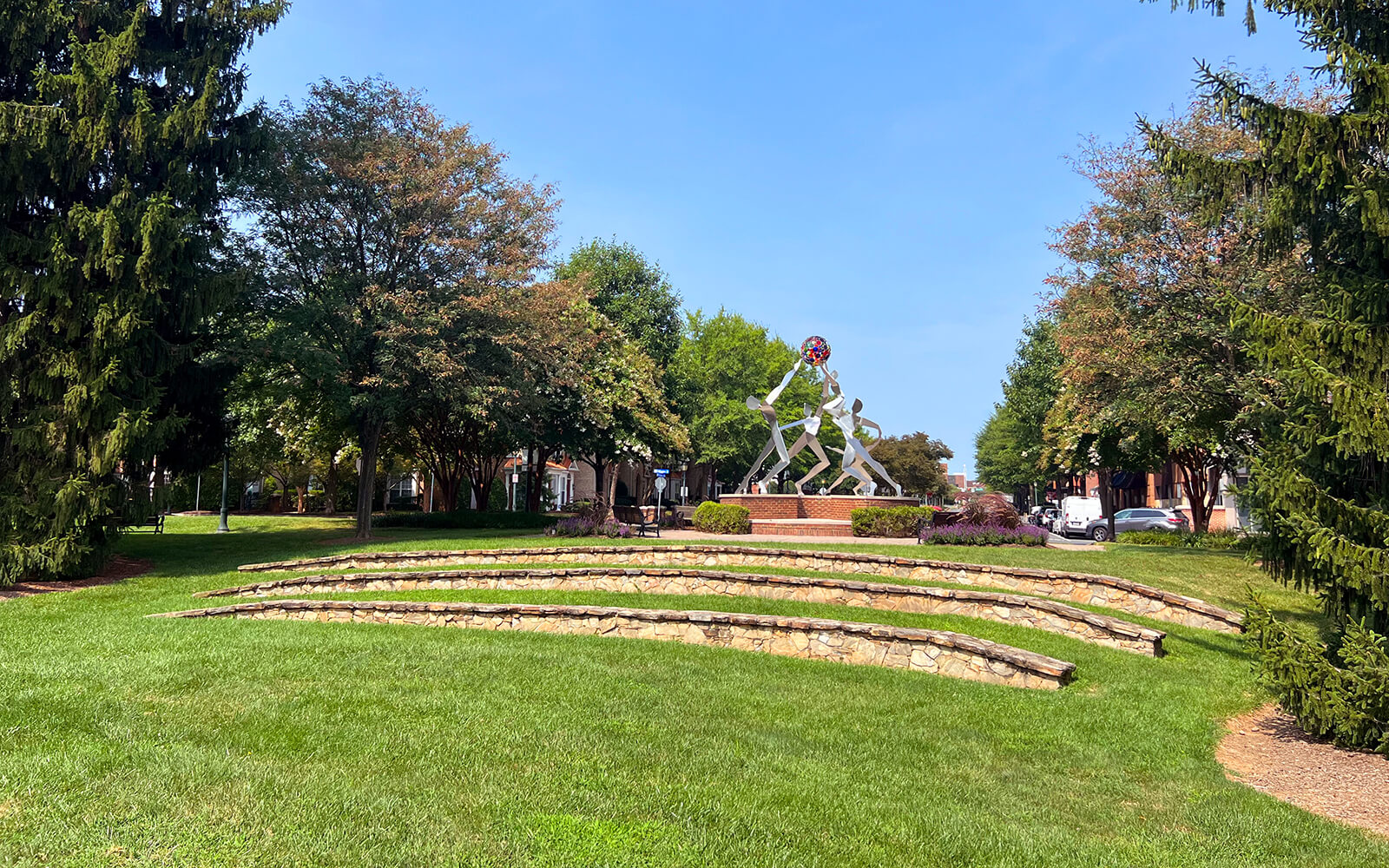 A tiered stone amphitheater faces a metal sculpture of human figures on a circular brick platform, surrounded by trees and green grass under a clear sky.