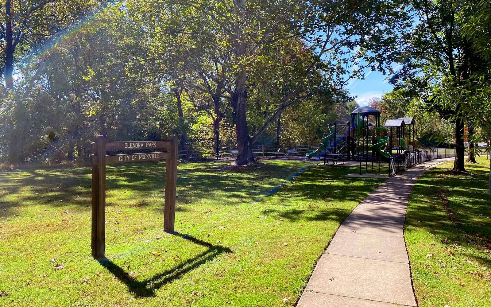A park with a wooden sign reading “Blenheim Park City of Rockville,” a playground structure, trees, and a paved path on a sunny day.