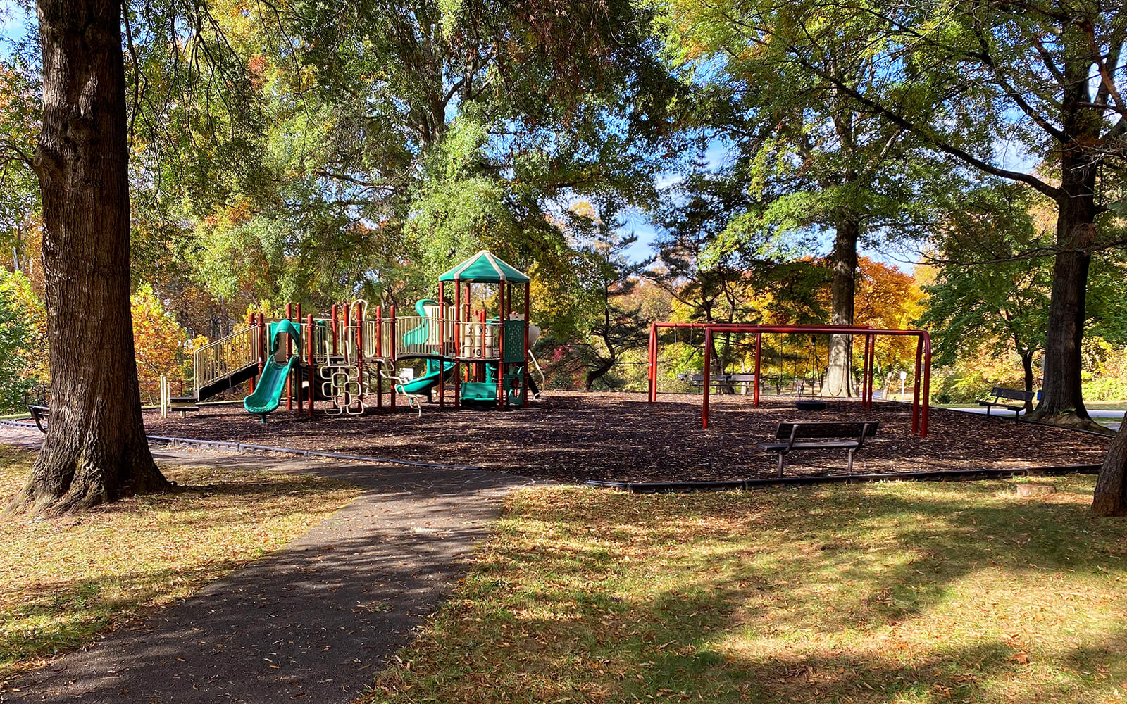 A playground with slides, climbing structures, and empty swings is surrounded by trees and shaded benches on a sunny day.