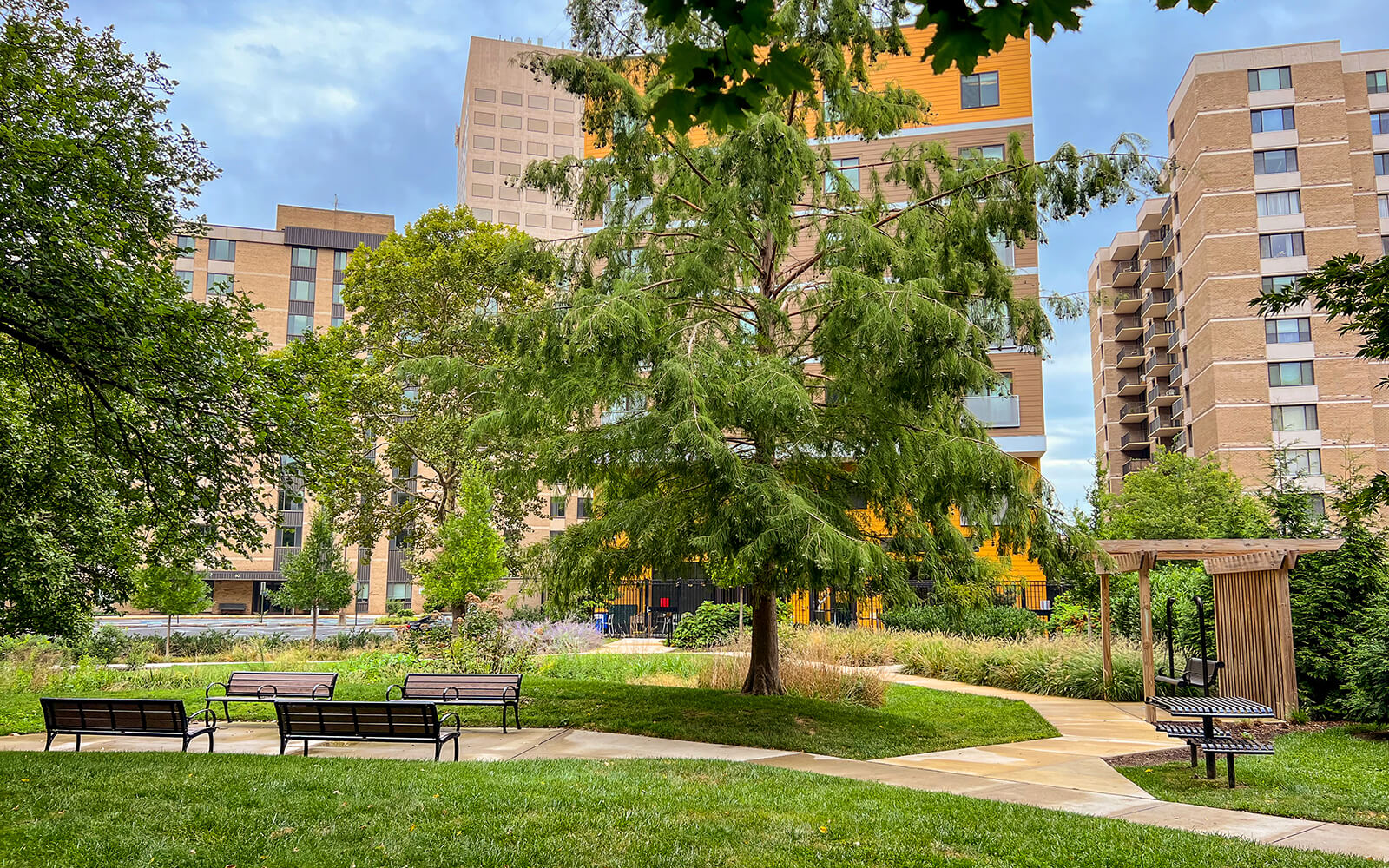 A landscaped urban park with benches and a pathway, surrounded by tall apartment buildings and trees under a partly cloudy sky.