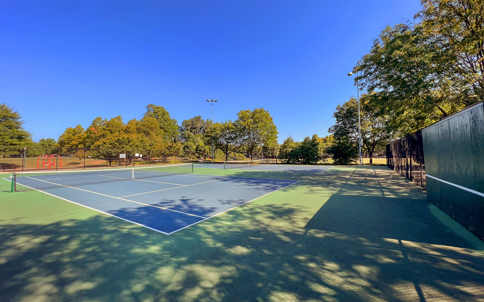 A tennis court in a park is surrounded by trees under a clear blue sky, with sunlight casting shadows on the court.