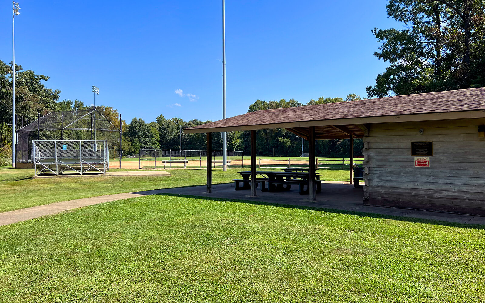 A covered picnic area with picnic tables sits next to a baseball field and backstop on a sunny day in a park.