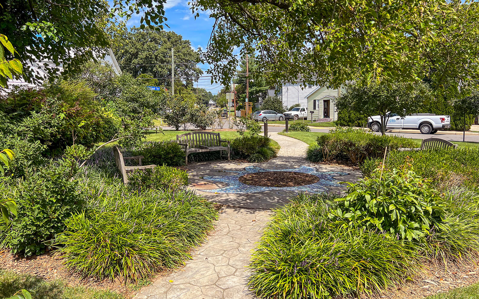 Small landscaped garden with benches, stone pathways, and surrounding greenery; houses, trees, and parked vehicles visible in the background.
