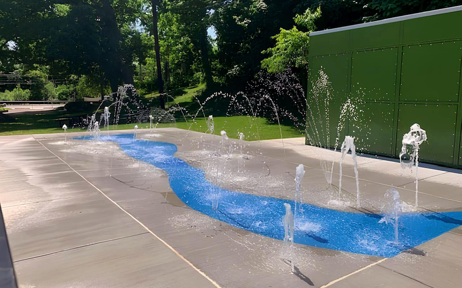 A splash pad with multiple water jets spraying over a blue, river-shaped design on a concrete surface, surrounded by grass and trees.