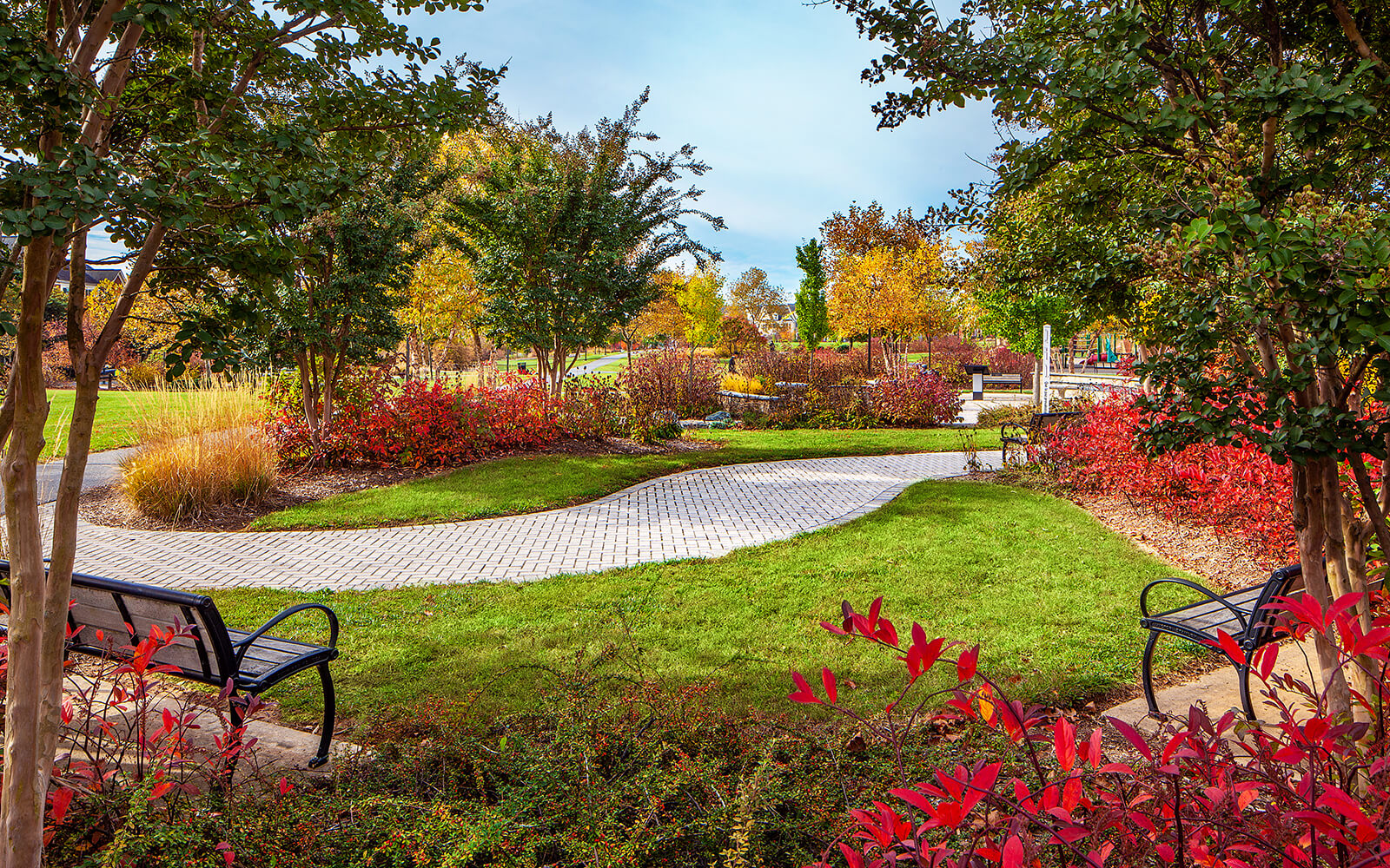 Curved stone path runs through a landscaped park with benches, green grass, and trees displaying autumn foliage under a partly cloudy sky.