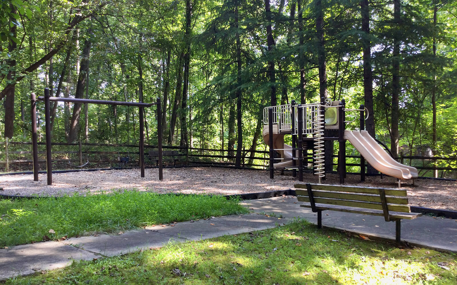 Empty playground in a wooded area with swings, a slide, climbing equipment, and a wooden bench. Sunlight filters through the trees.