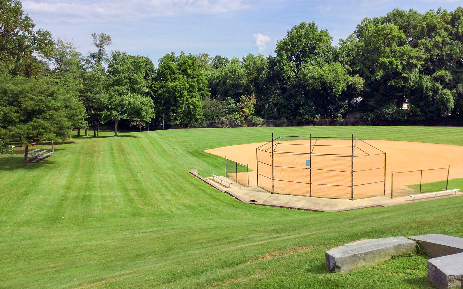 A baseball field with a dirt infield and fenced backstop is surrounded by green grass and trees under a partly cloudy sky.