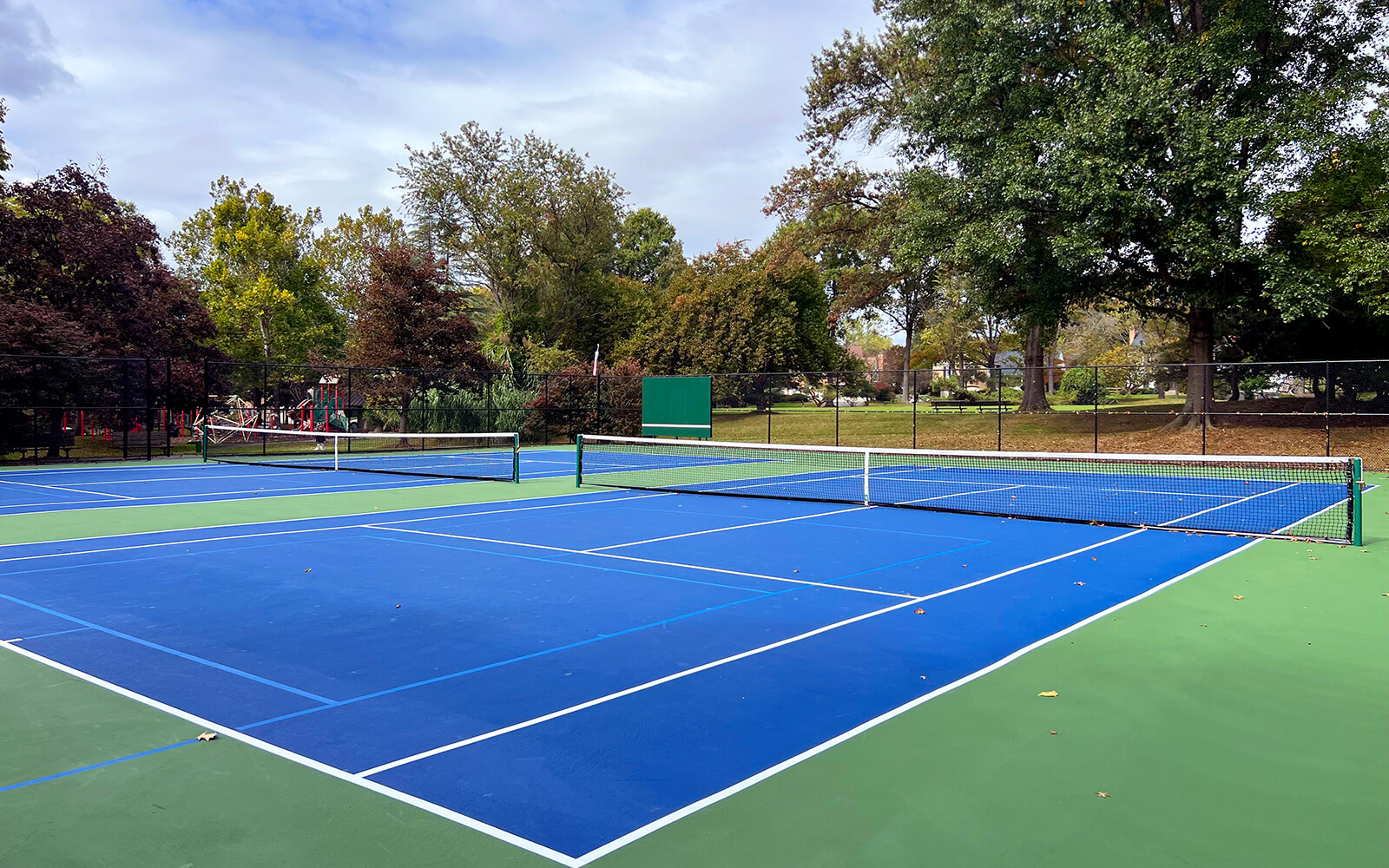 A blue and green outdoor tennis court is shown empty, surrounded by trees and a chain-link fence on a cloudy day.