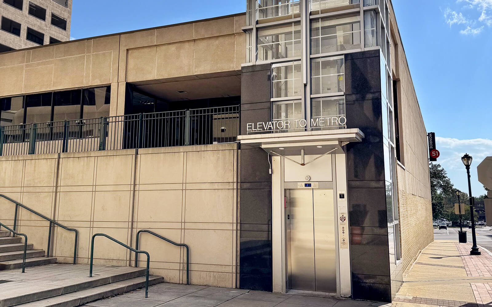 Street-level view of a modern elevator entrance labeled "Elevator to Metro" next to a beige building, with stairs and a railing on the left.