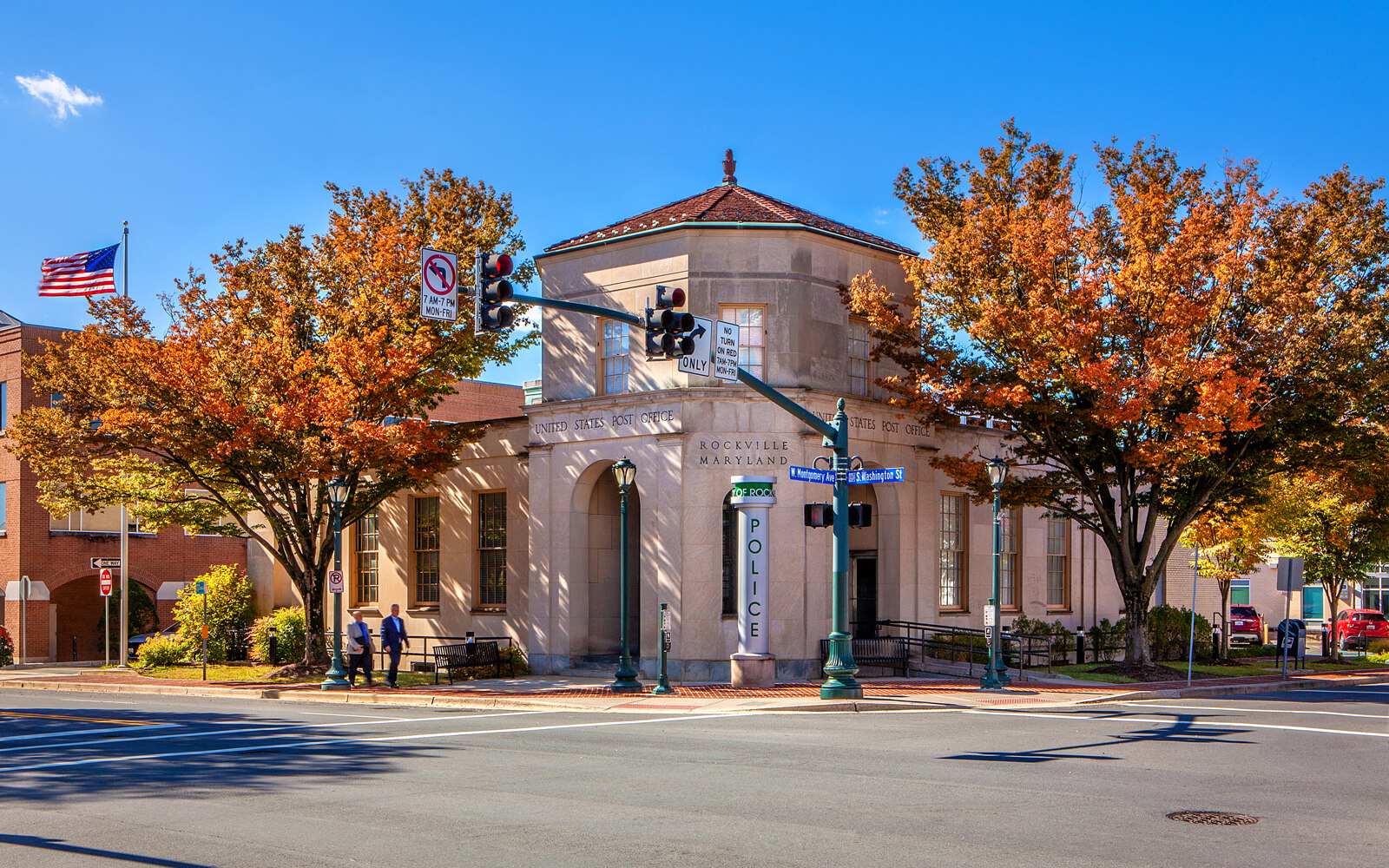 A small police station building with autumn trees in front, located at a street intersection with visible traffic lights and a U.S. flag in the background.
