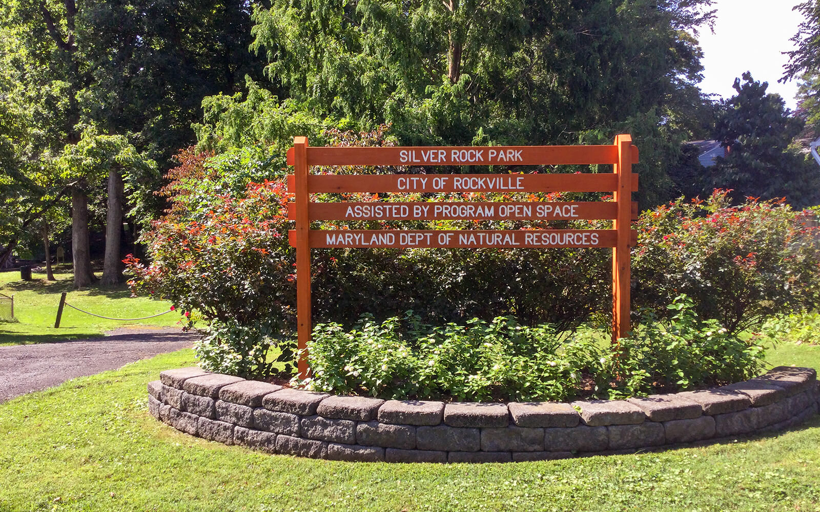 An orange wooden sign at Silver Rock Park in Rockville stands on a stone-bordered garden, surrounded by bushes and trees.