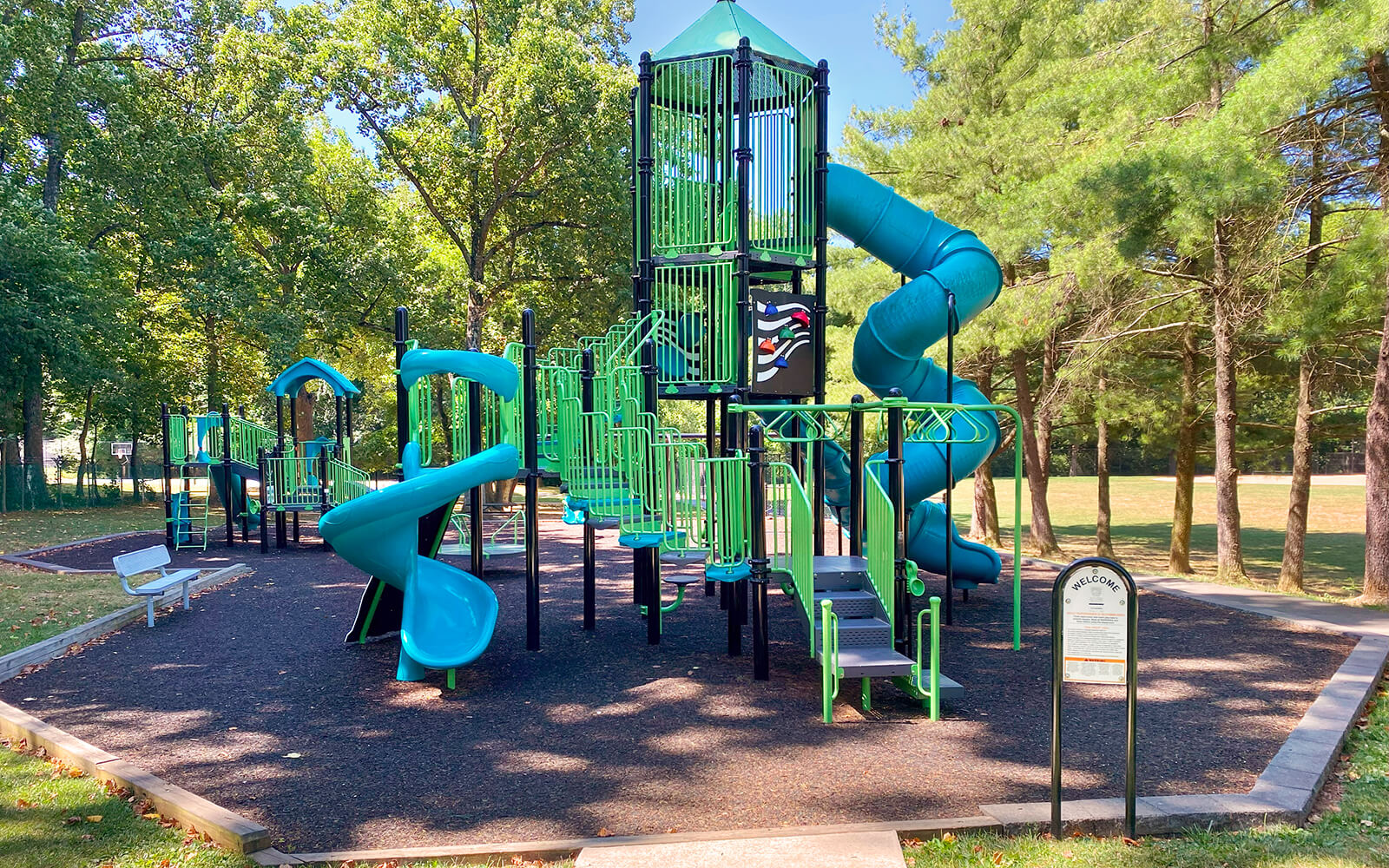 A modern playground with green and blue climbing structures, slides, and a spiral tube slide, surrounded by trees and benches on a sunny day.