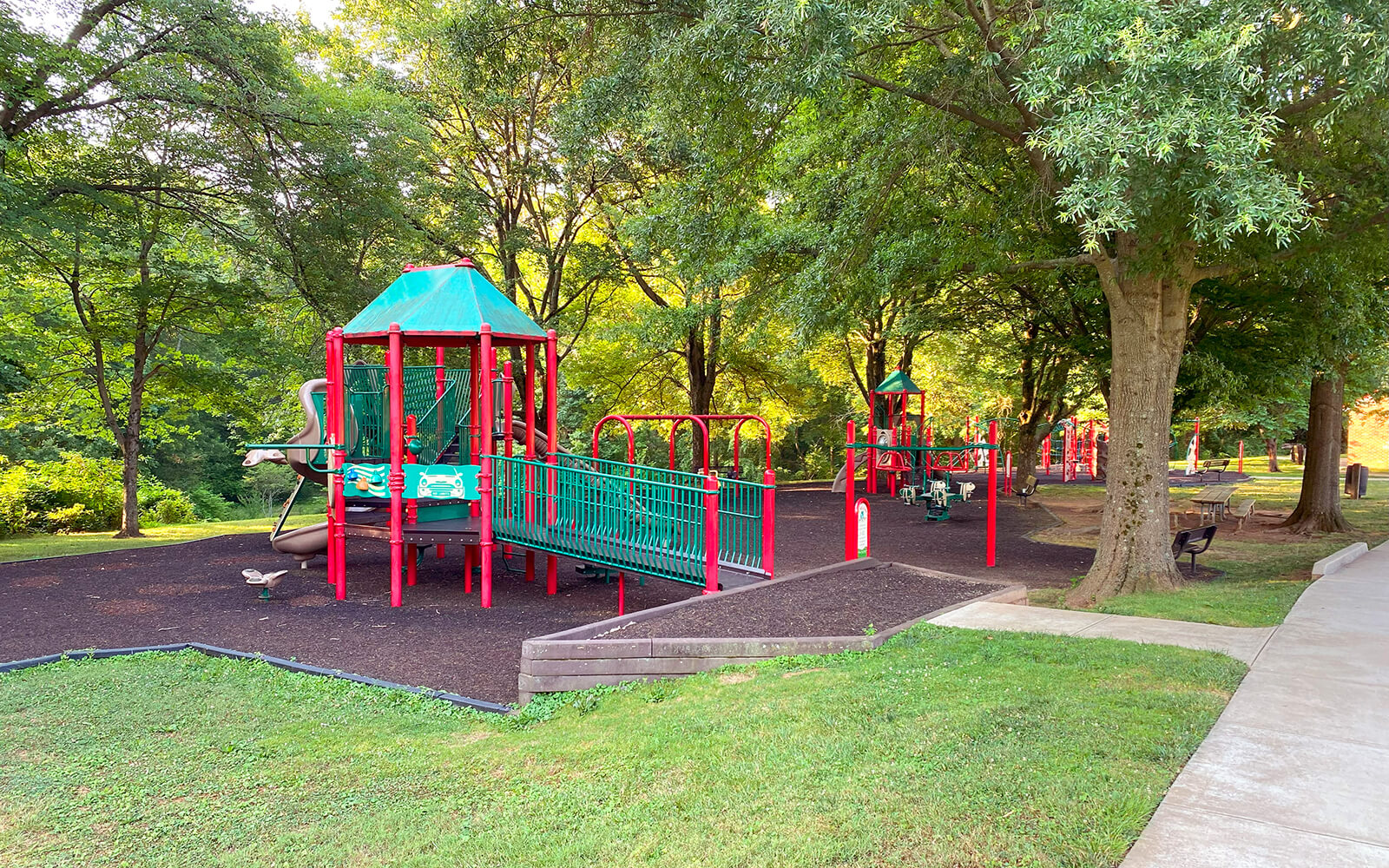 A playground with red and green equipment, including slides and bridges, is surrounded by grass and trees on a sunny day.