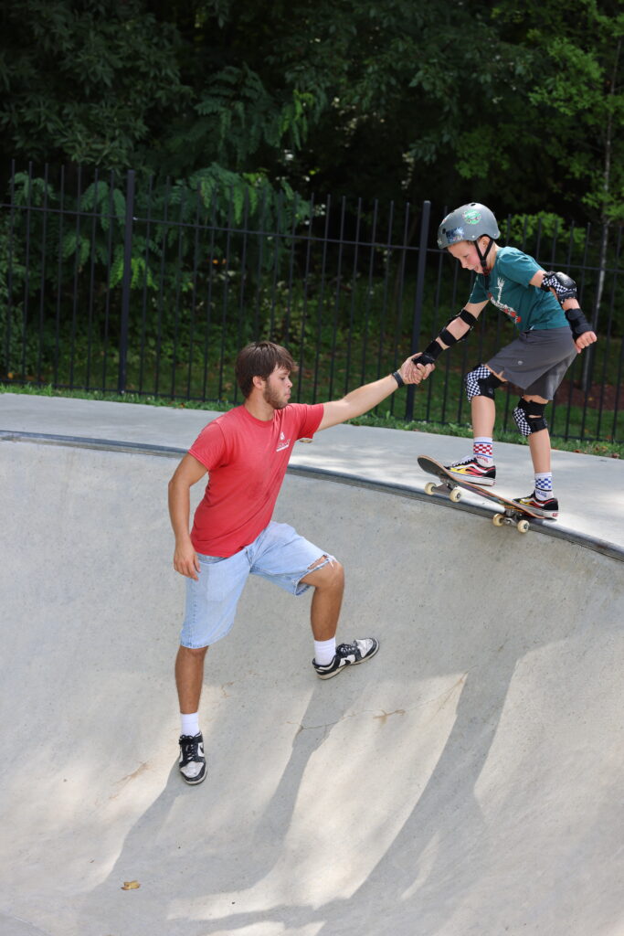 An adult helps a child on a skateboard at a skate park, holding their hand for support near the edge of a concrete bowl.