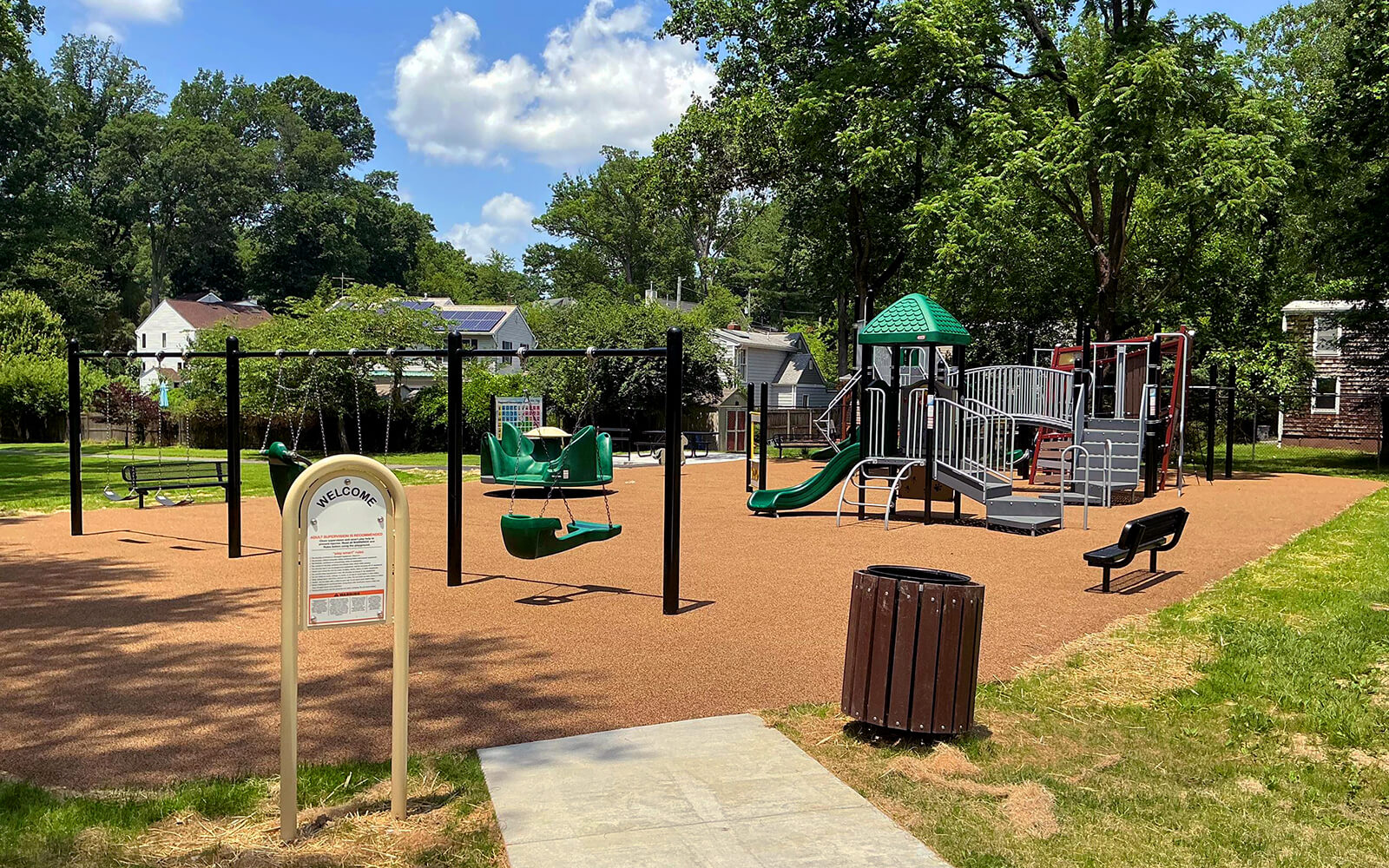 A playground with swings, slides, climbing structures, benches, and a trash can, surrounded by trees and nearby houses under a partly cloudy sky.