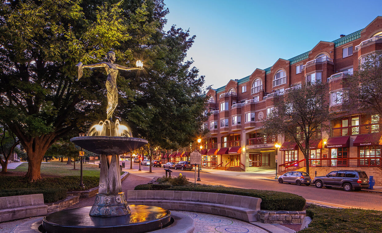 A bronze fountain statue in a small plaza is surrounded by trees and brick buildings with shops and apartments at dusk.