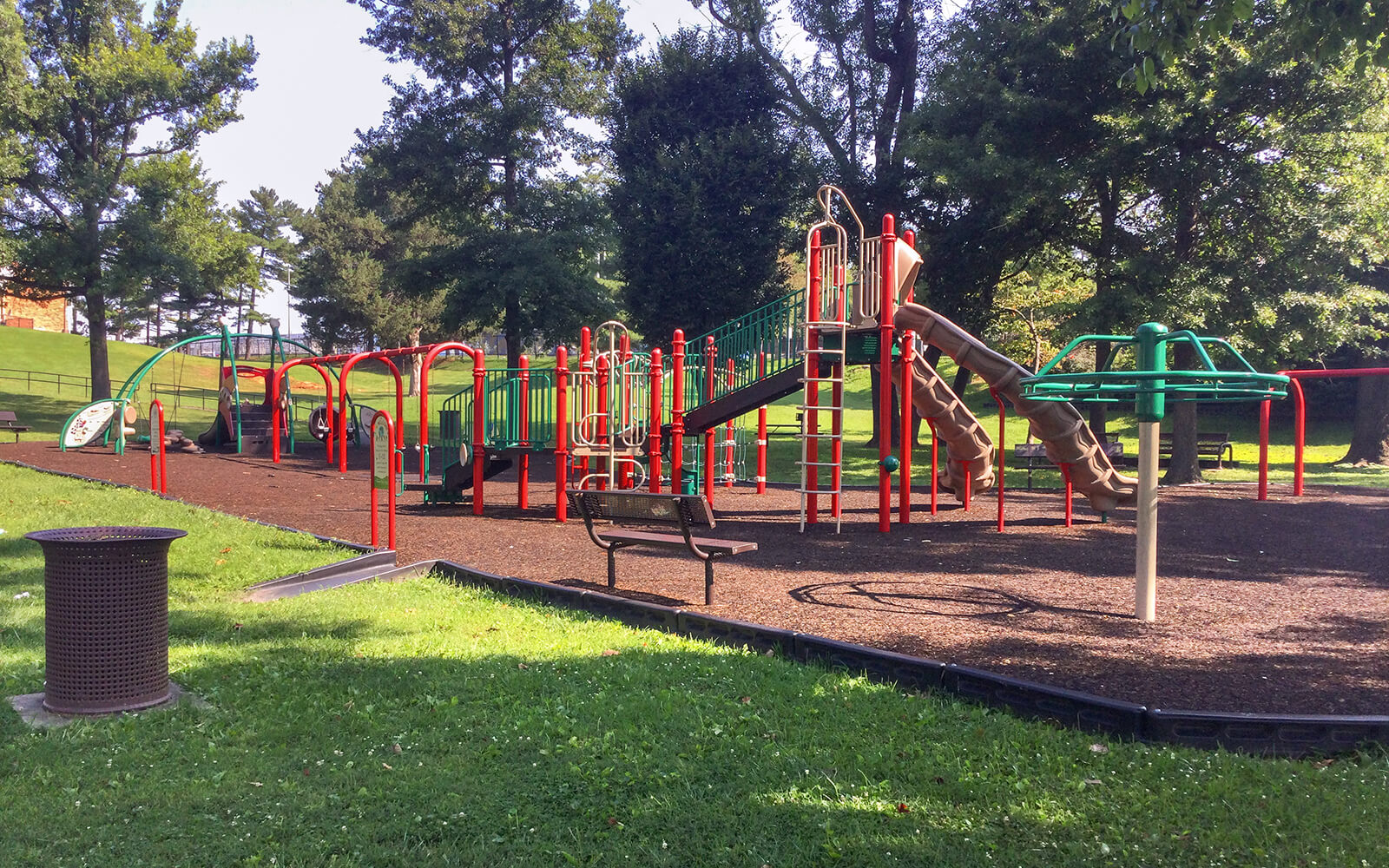 Playground with slides, climbing structures, and monkey bars surrounded by grass and trees; a bench and trash can are in the foreground.