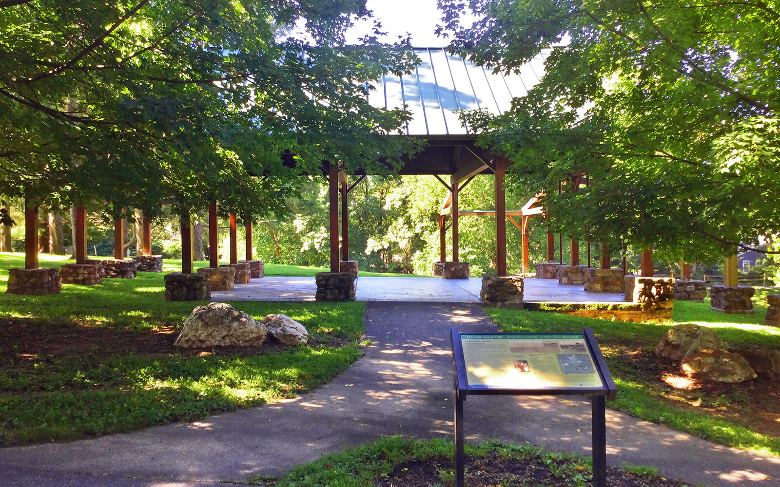 A shaded pavilion with a metal roof stands in a green park, surrounded by trees, stone posts, and a paved pathway with an informational sign in the foreground.