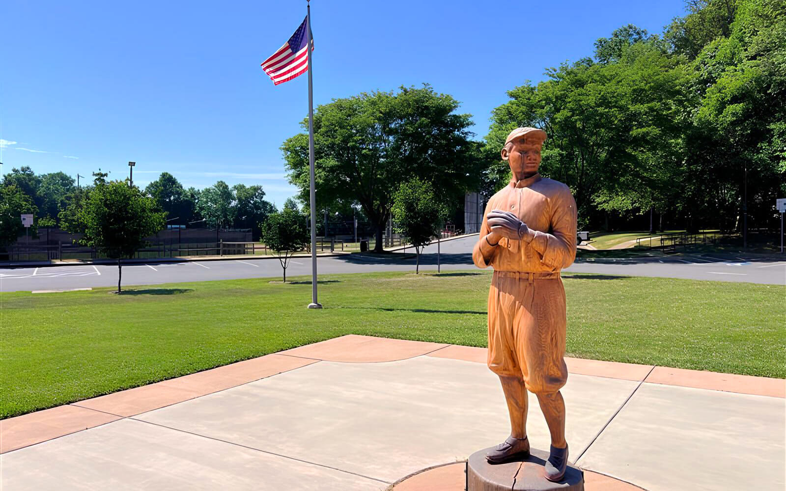 A statue of a baseball player wearing vintage uniform stands on a paved area, with an American flag and trees in the background.