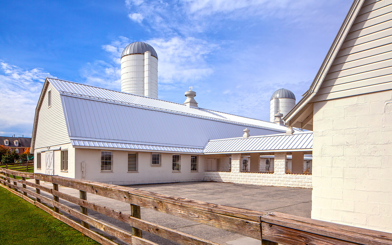 White barn buildings with metal roofs and silos, surrounded by a wooden fence under a partly cloudy blue sky.