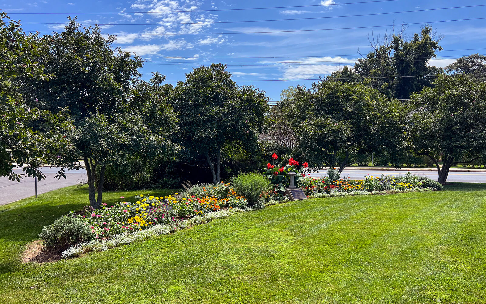 A landscaped garden bed with colorful flowers and green shrubs beside a lawn, bordered by trees under a partly cloudy blue sky.