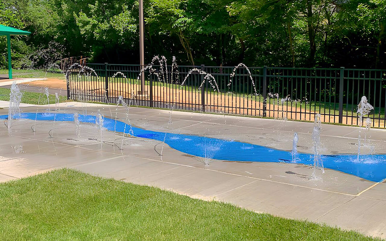 A splash pad features several water jets spraying arcs over a blue-painted section of concrete, with a fence and trees in the background.