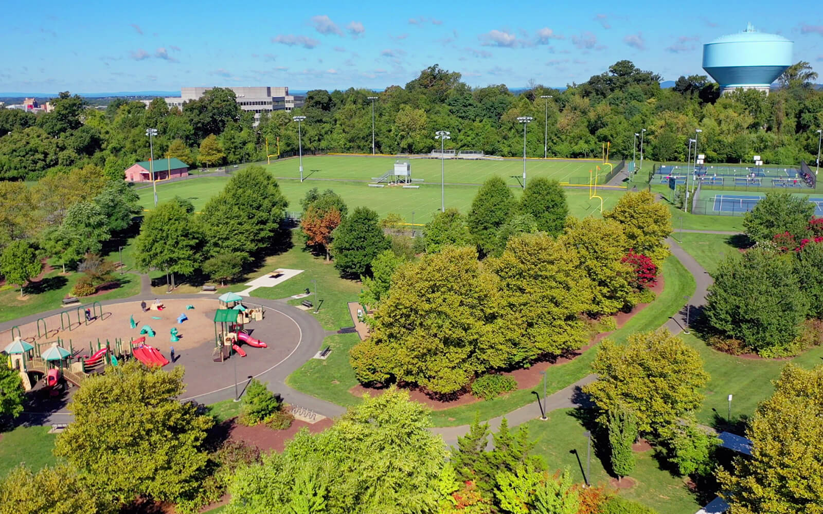 Aerial view of a park with a playground, sports fields, tennis courts, walking paths, trees, and a water tower in the background.