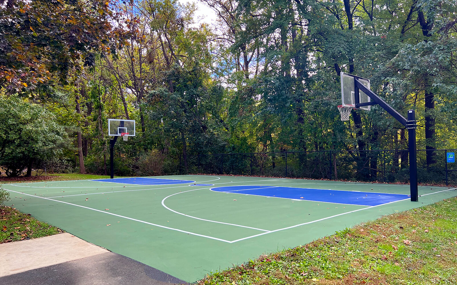 An outdoor basketball court with two hoops is surrounded by trees and greenery on a clear day.