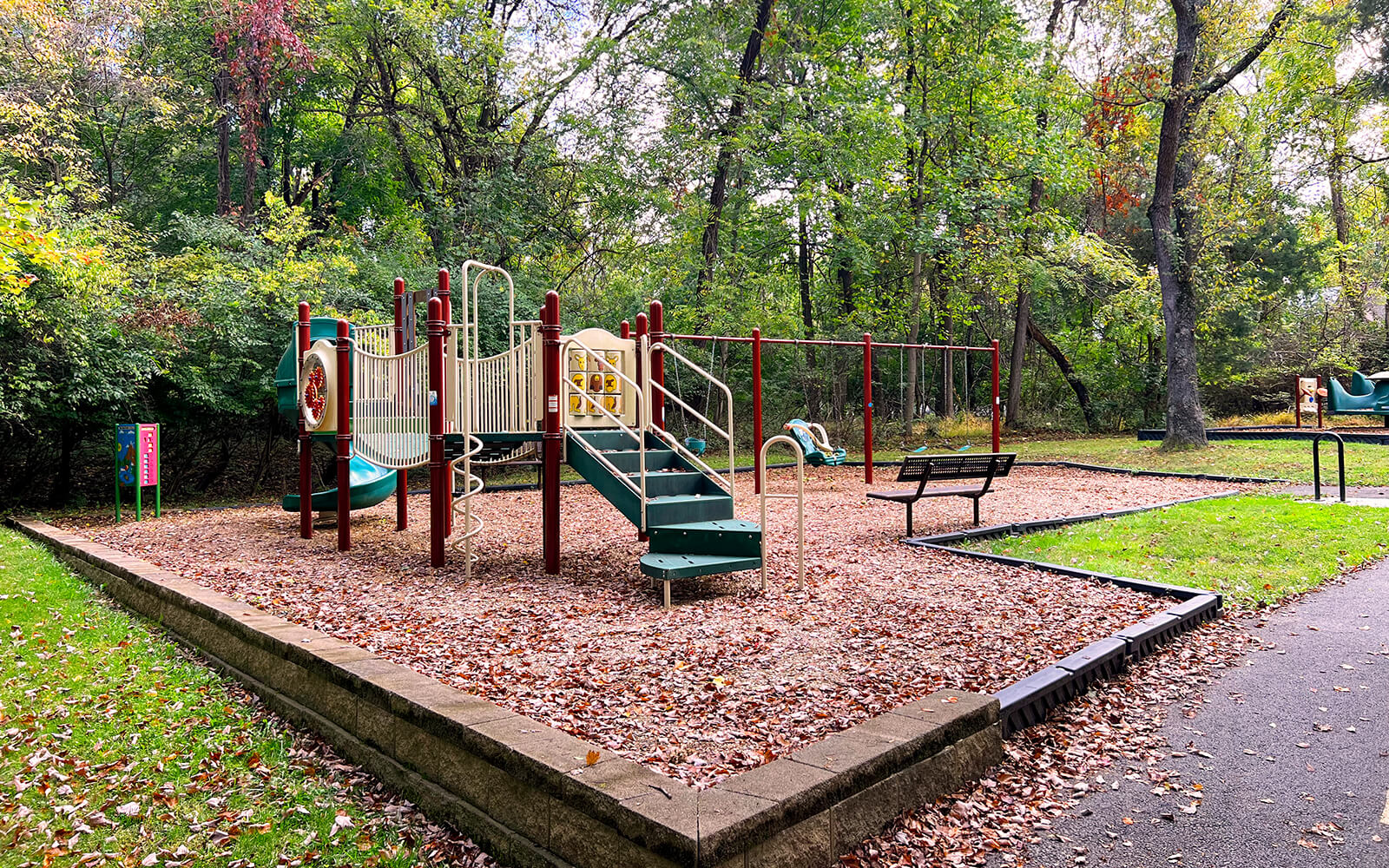 A playground with slides, climbing equipment, and benches is surrounded by trees and fallen leaves on the ground.