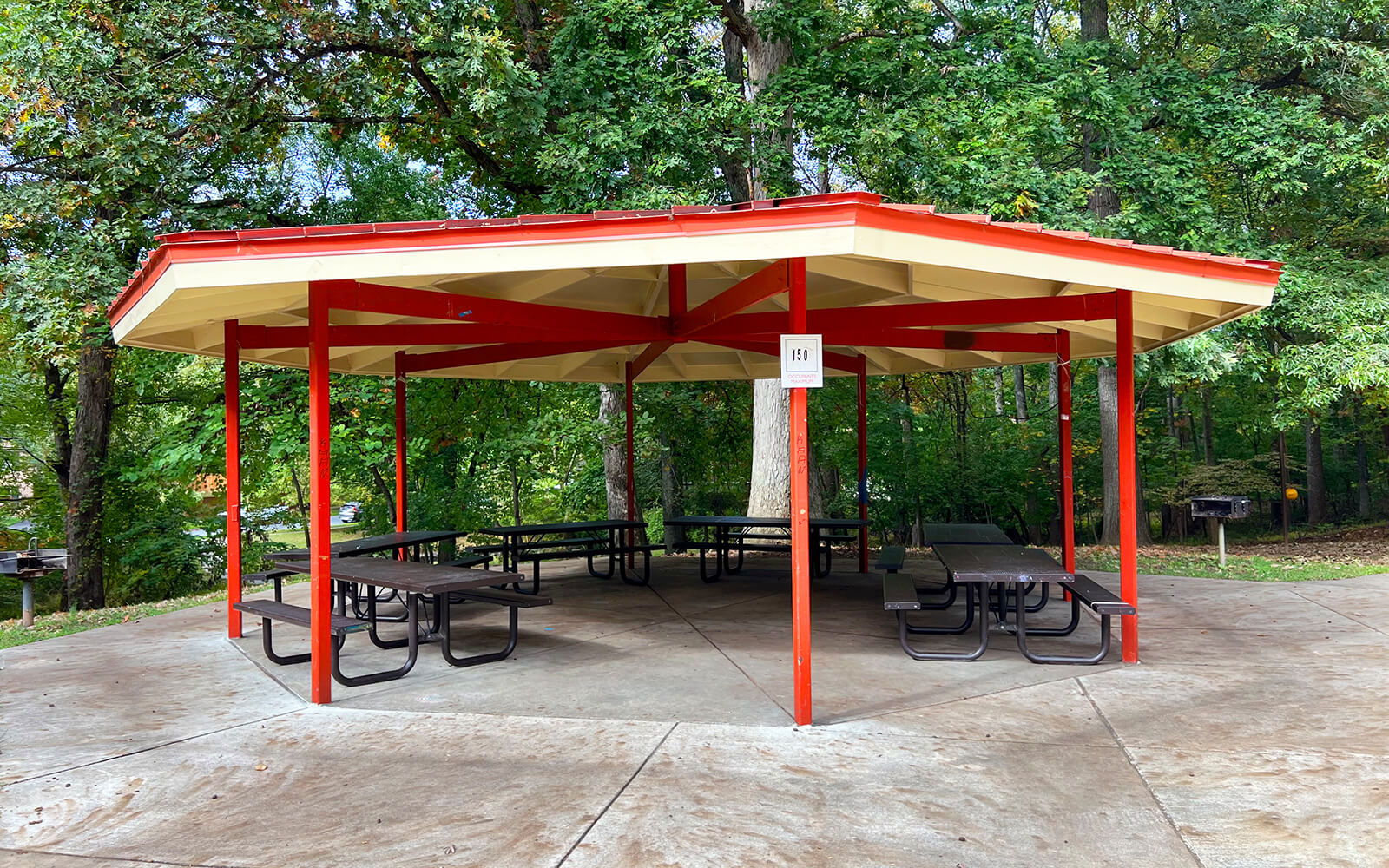 Hexagonal pavilion with a red roof and red support beams, containing several picnic tables, set in a wooded park area.