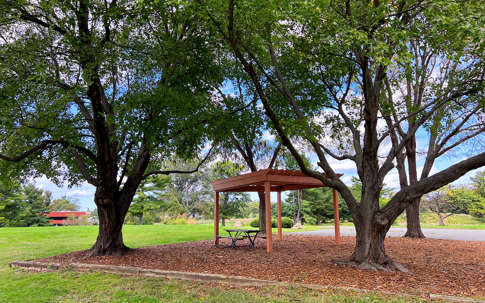 A small wooden pavilion with a picnic table sits under large trees in a park, surrounded by grass and mulch on a partly cloudy day.