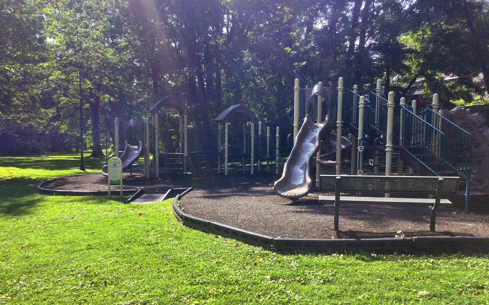 A playground with slides, climbing structures, and a bench is situated in a grassy, tree-filled park on a sunny day.