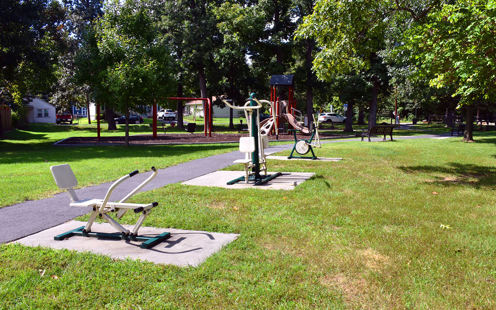 Outdoor fitness equipment is installed along a paved path in a grassy park, with trees and playground equipment visible in the background.