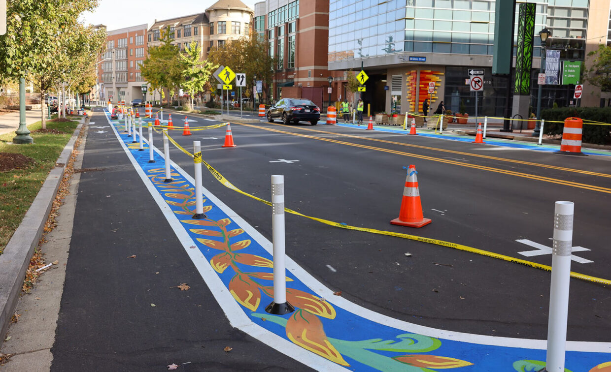 A city street with a newly painted bike lane featuring colorful designs, separated by white posts and orange traffic cones, with buildings and caution tape in the background.