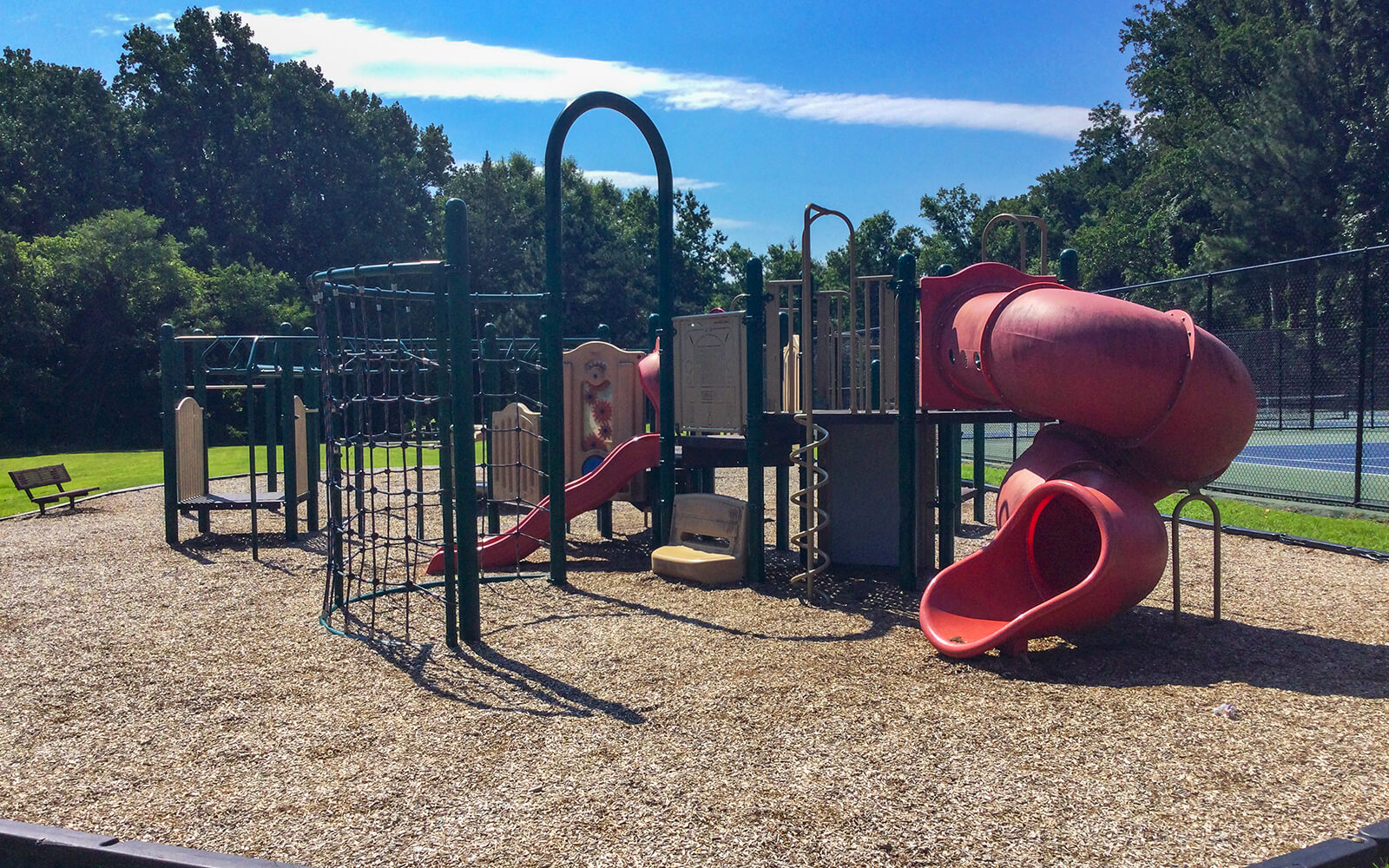 Outdoor playground with slides, climbing structures, and tunnels on a mulch surface, surrounded by trees and a tennis court in the background on a sunny day.