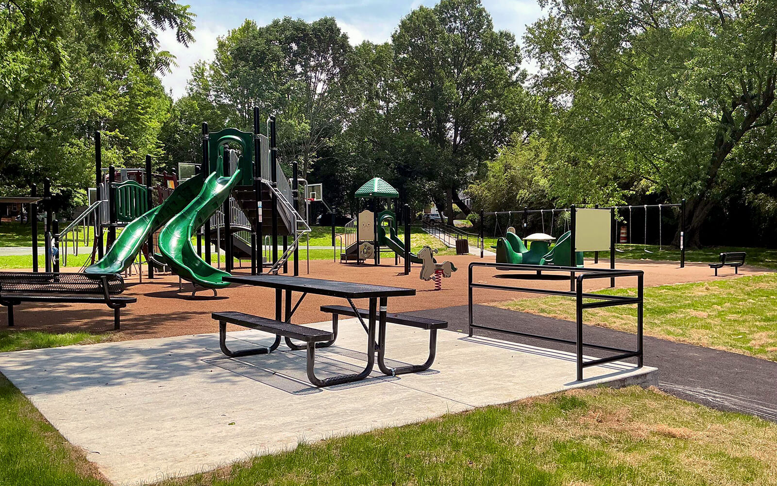 Playground with green slides, climbing structures, picnic tables, benches, and rubber flooring, surrounded by trees on a sunny day.