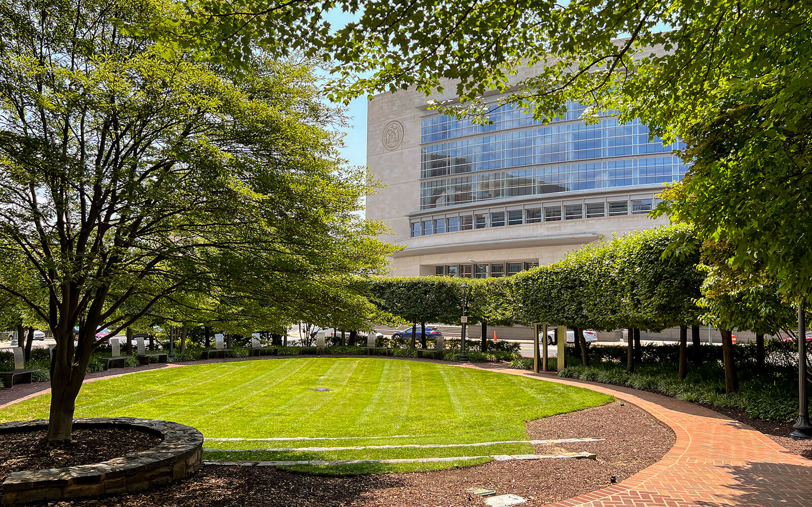A circular lawn bordered by trees and a brick path, with a large modern glass building in the background on a sunny day.