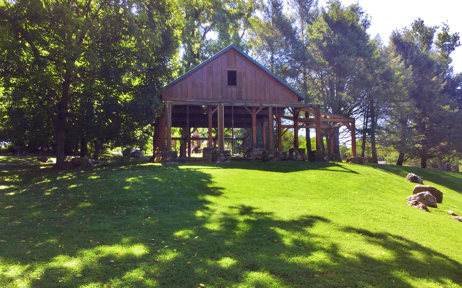 A wooden pavilion with open sides stands on a grassy lawn surrounded by trees, with sunlight filtering through the foliage.