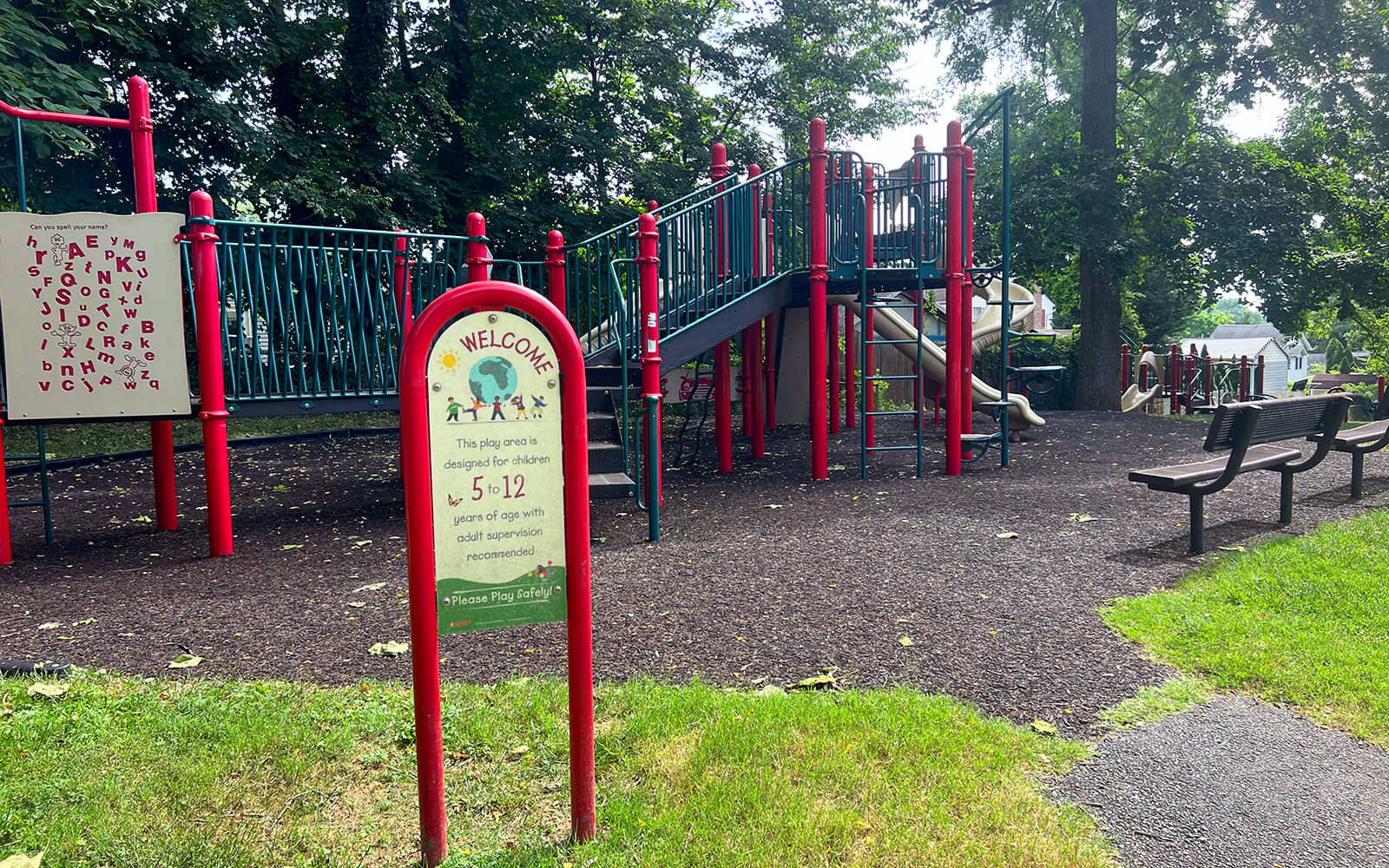 A playground with red and green equipment, a slide, climbing features, benches, and a sign stating it is designed for children ages 5-12. Trees and grass surround the area.