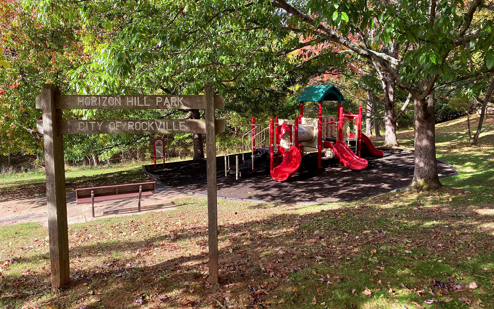 A wooden sign for Horizon Hill Park stands near a playground with red slides and a green canopy, surrounded by trees and grass.