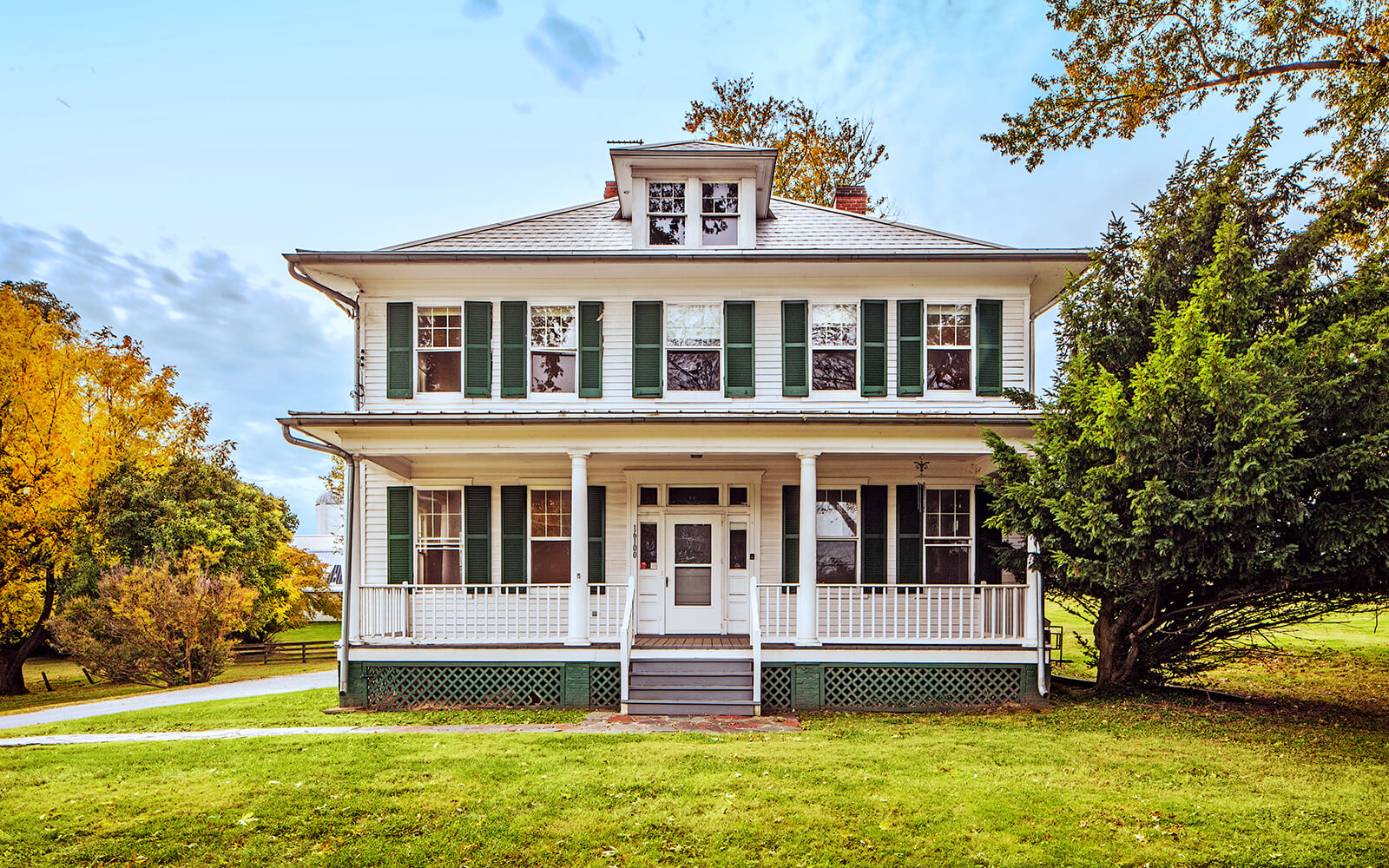 A two-story white house with green shutters, a covered front porch, and surrounding trees, photographed on a clear day.