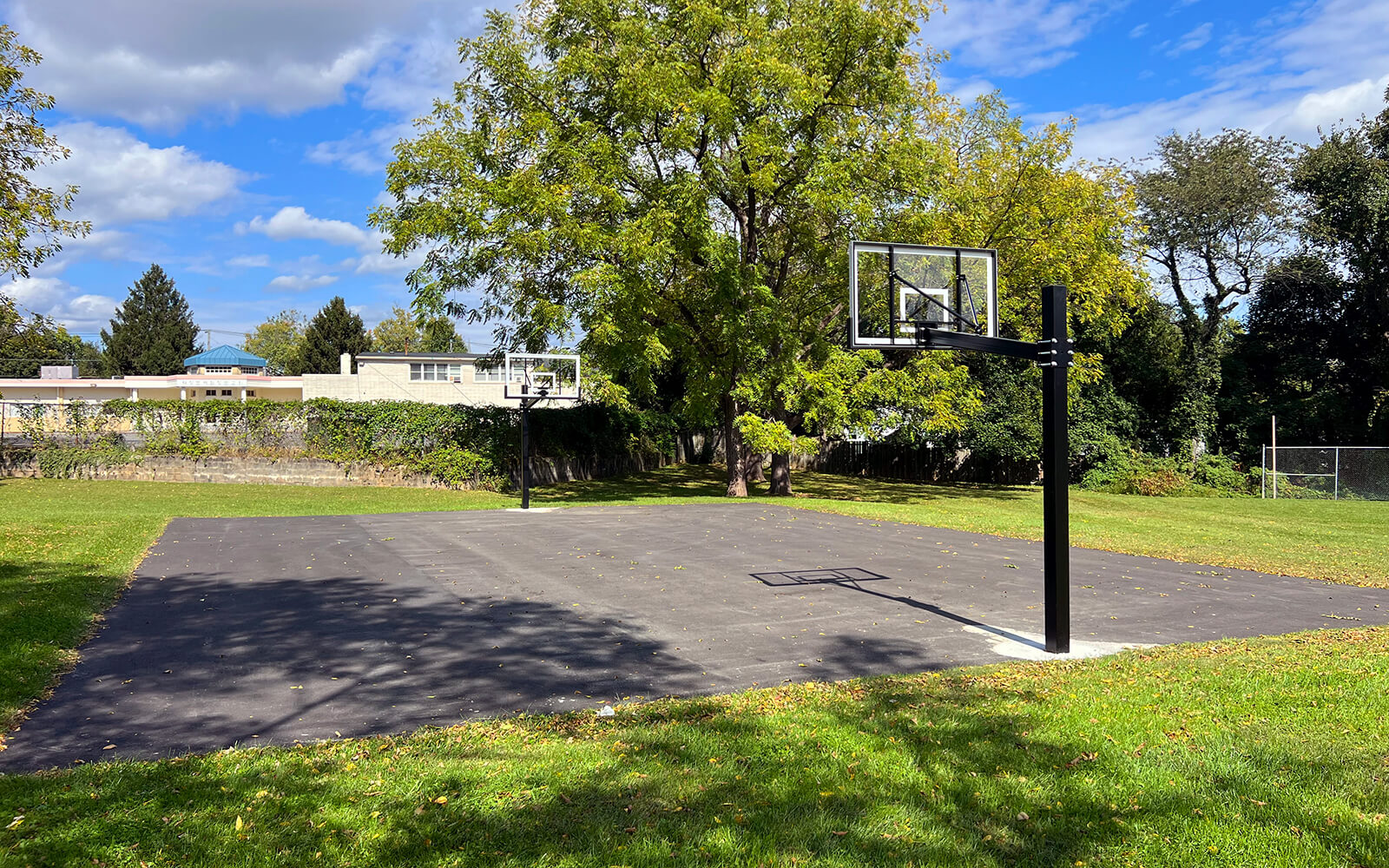 Outdoor basketball court with two hoops, surrounded by grass and trees, under a partly cloudy sky. Some buildings are visible in the background.