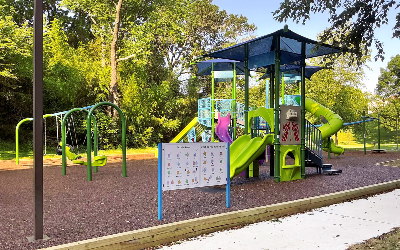 Outdoor playground with green and blue slides, swings, climbing structures, and a posted rules sign, surrounded by trees and set on a mulch surface.