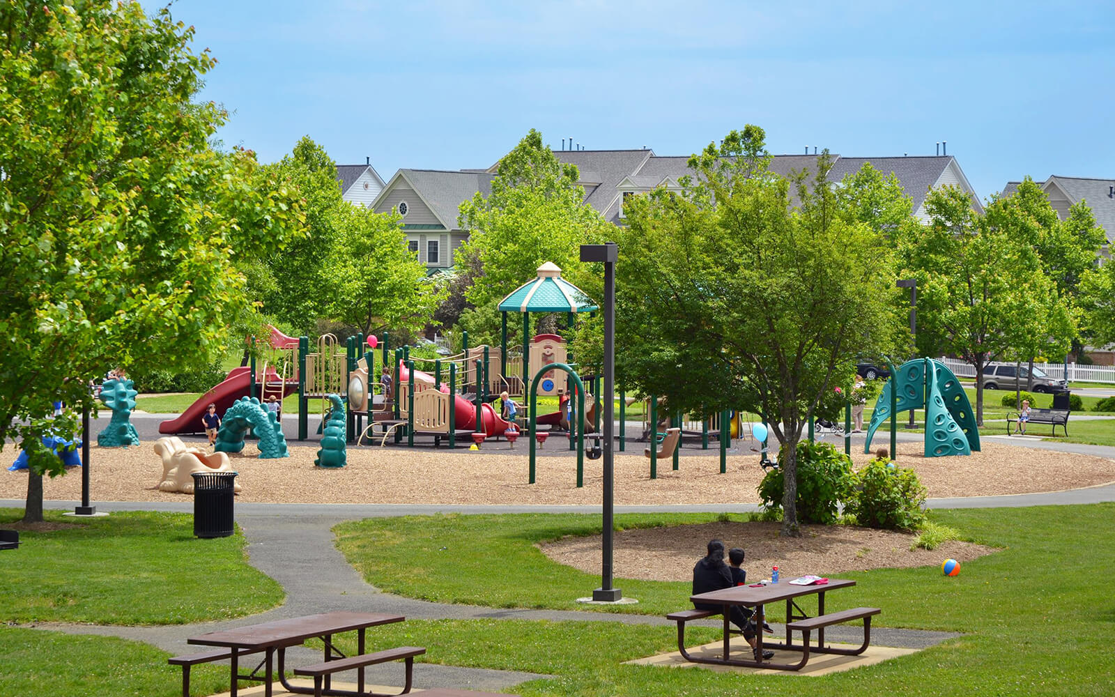 A playground with slides and climbing structures is surrounded by trees and nearby houses. Two people sit at a picnic table in the foreground.
