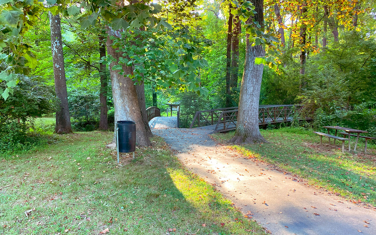 A paved path leads to a small metal footbridge in a wooded park area, with a trash bin and picnic table nearby.
