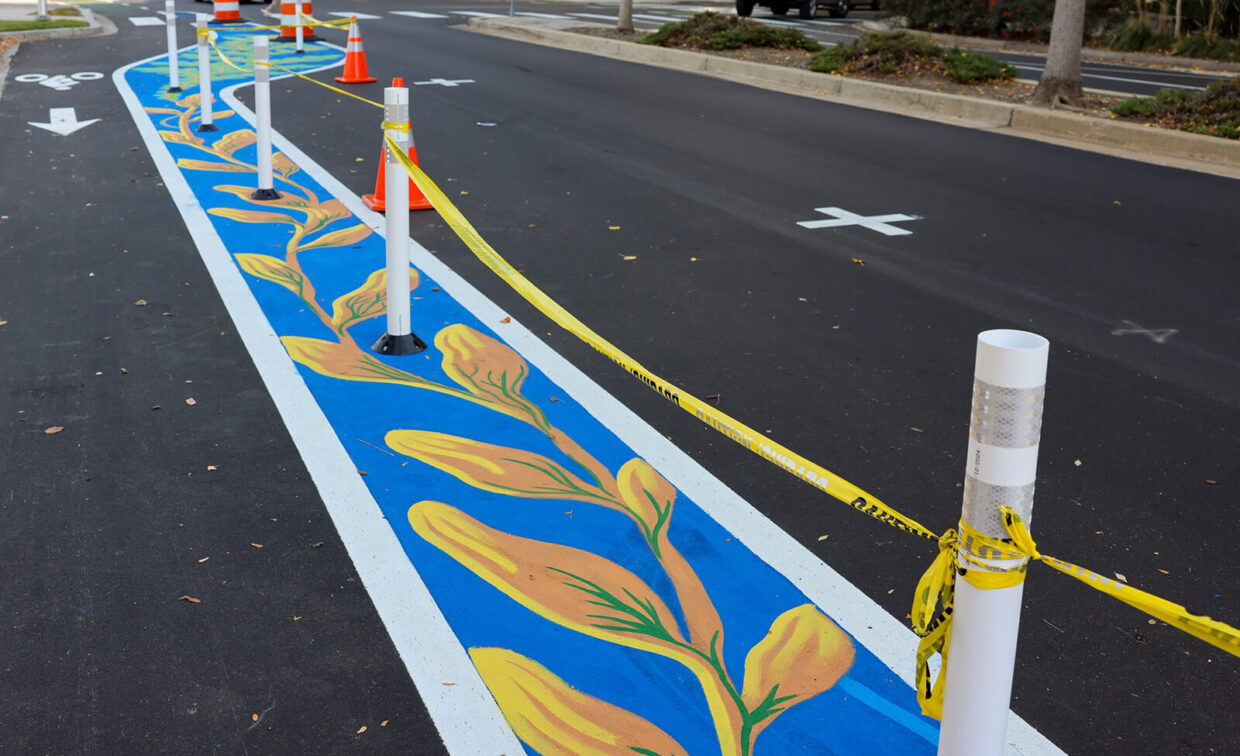 A street with a blue bike lane decorated with yellow flower designs, bordered by white poles and yellow caution tape, with orange traffic cones nearby.