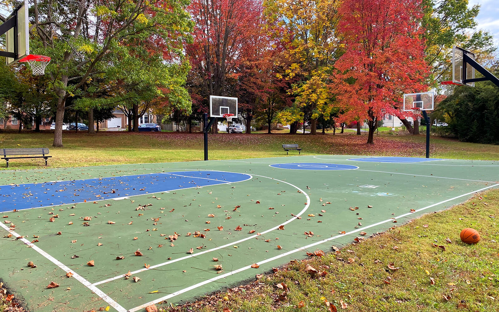 Outdoor basketball court with three hoops, scattered autumn leaves on the ground, and a basketball resting near the edge of the court. Trees with fall foliage are in the background.