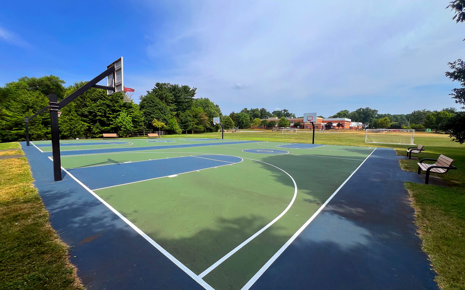 Outdoor basketball court with three hoops, surrounded by green grass, trees, benches, and a soccer goal in the background under a clear sky.